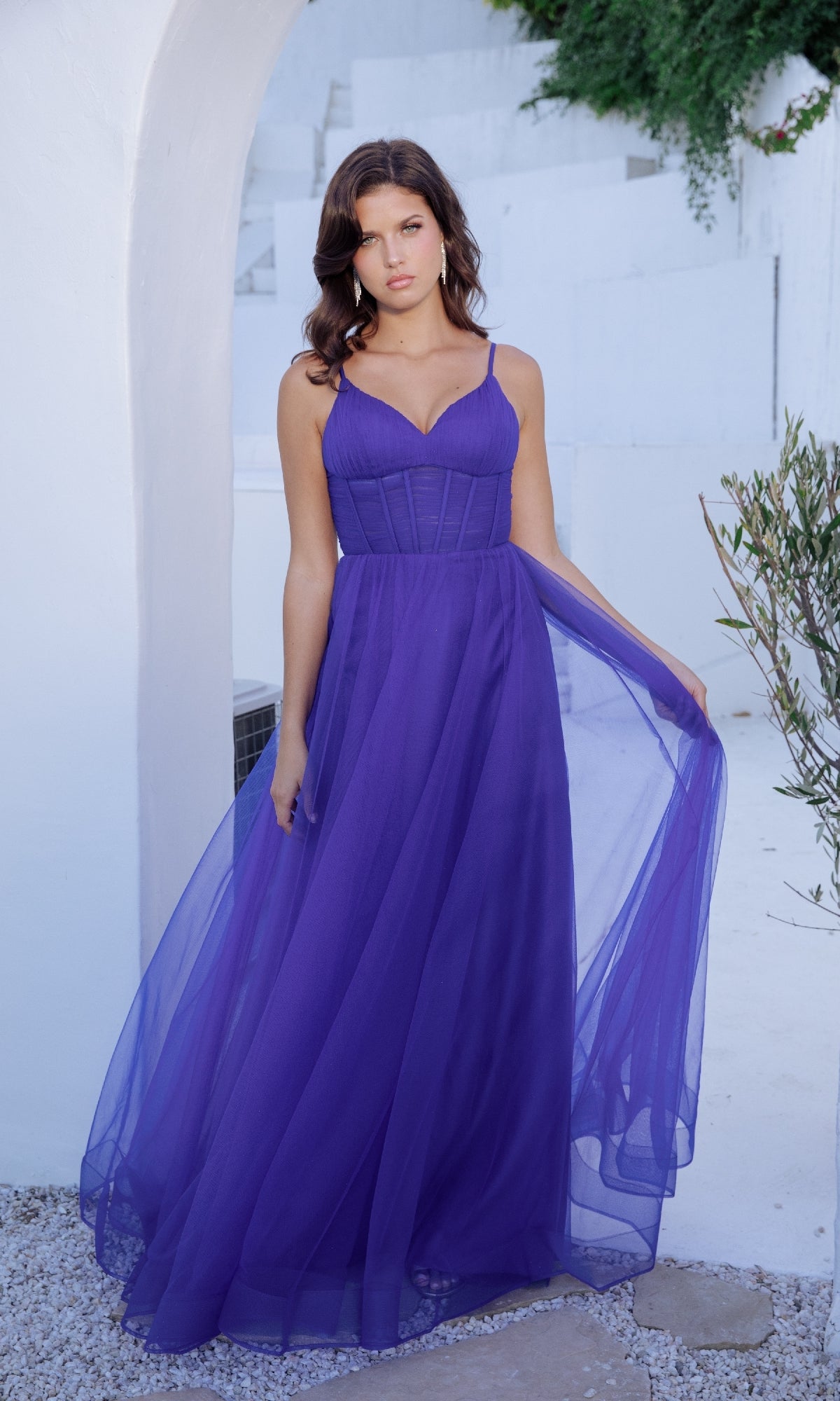 A woman stands outdoors before a white wall, wearing the Corset Long Tulle Prom Ball Gown: Eureka 9177—a sleeveless, floor-length purple dress with a fitted corset bodice and sheer flowing skirt.